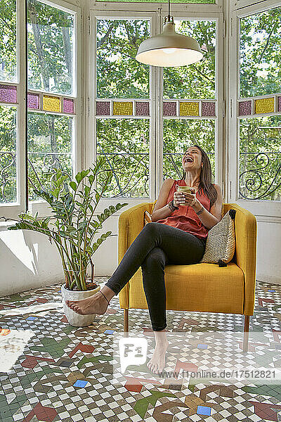 Fröhliche Frau mit Tasse sitzt zu Hause auf einem Sessel am Fenster