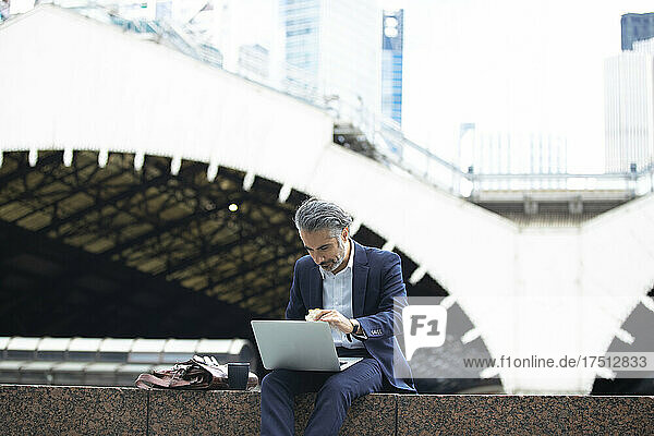 Businessman using laptop while sitting on retaining wall in city