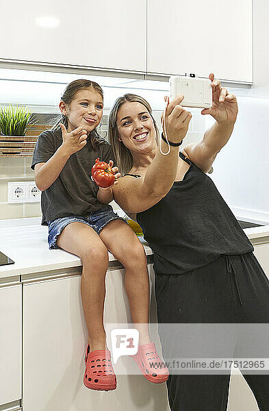 Happy mother taking selfie with daughter eating tomato on kitchen counter at home