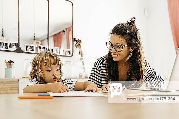 Smiling mother looking at cute daughter painting on dining table at home