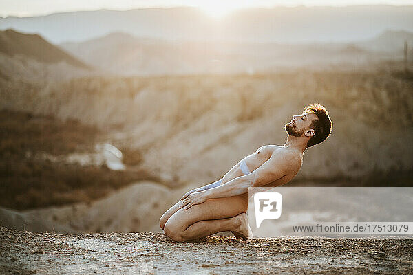 Naked mid adult man kneeling on land at desert during sunset