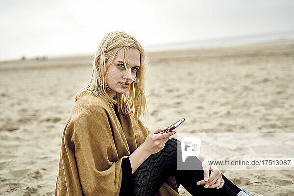 Netherlands  portrait of blond young woman with smartphone sitting on the beach