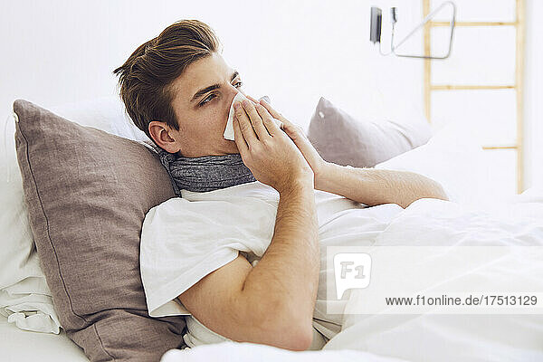 Sick young man blowing nose while lying on bed at home