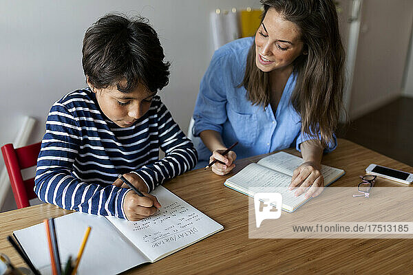 Young woman assisting boy in writing homework on table at home