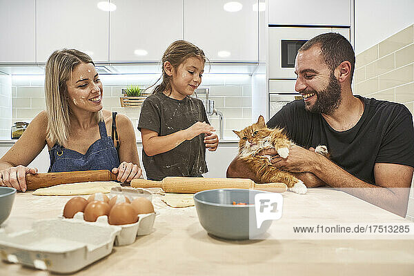 Mutter knetet Teig mit Nudelholz  während Vater und Tochter mit Katze auf dem Küchentisch spielen
