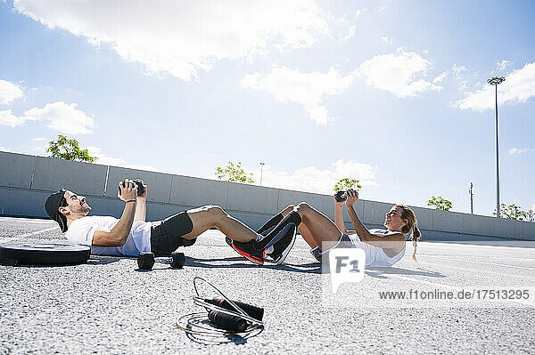 Couple holding dumbbells while lying on road against sky during sunny day