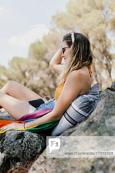 Young woman listening music through headphones while sitting with rainbow flag on rock