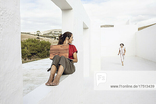 Mother using laptop with wooden cover  watching daughter running in white architecture