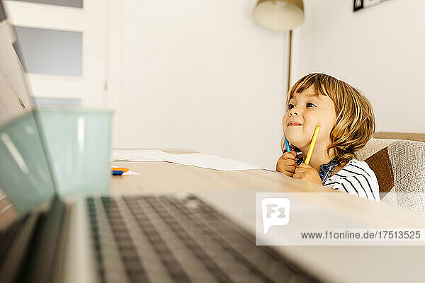 Cute girl holding crayons looking away while sitting at dining table