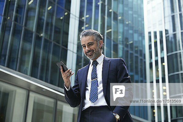 Lächelnder Geschäftsmann benutzt Mobiltelefon vor Bürogebäude in der Stadt