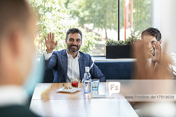 Smiling businessmen waving hands at coworker while sitting in restaurant