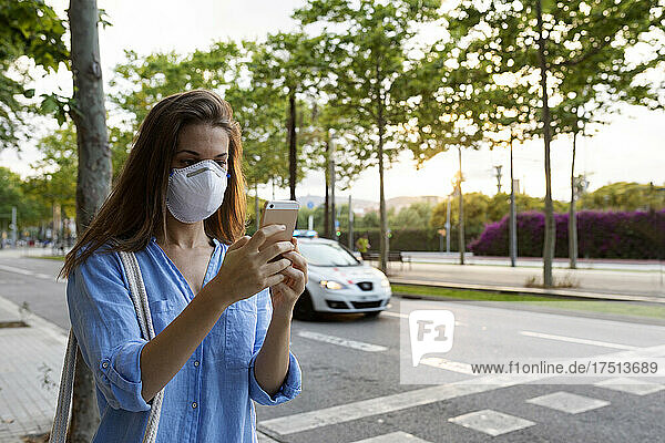 Young woman wearing mask using smart phone while standing on street in city