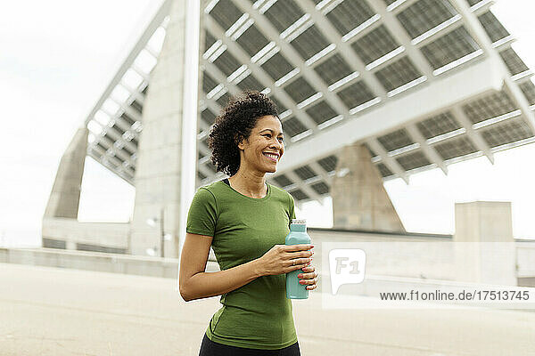Smiling woman holding water bottle looking away while standing against built structure in city