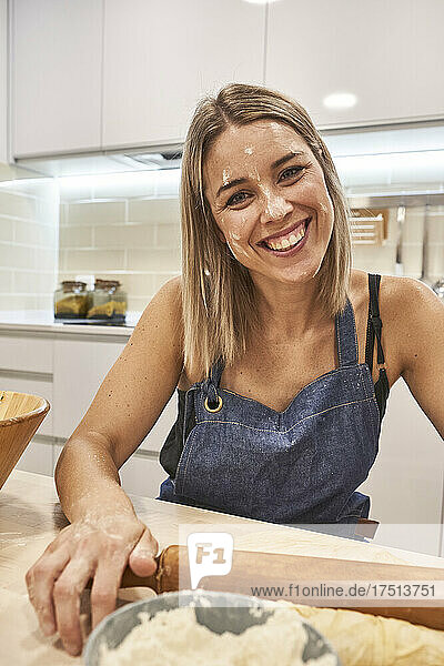 Close-up of smiling woman with flour on face holding rolling pin over table in kitchen