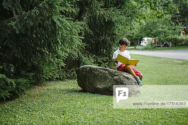 A child sits on boulder by side of road reading picture book