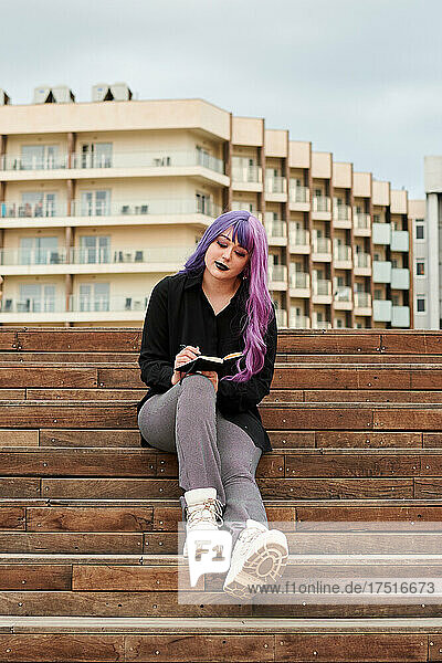 Non-binary person with purple hair writes in a notebook on the street