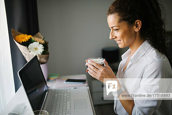 Woman looking at he laptop and smiling while holding a cup of co