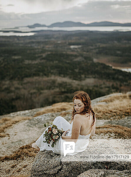 Bride in wedding gown sits on mountain with expansive view