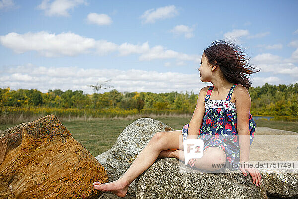 Small windblown girl with bare feet sits on boulder outside in summer