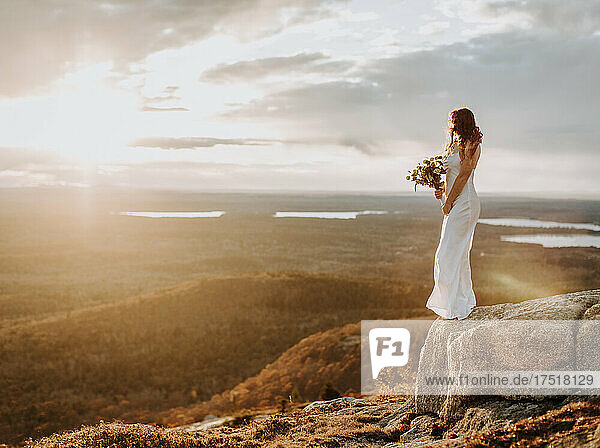 Bride wedding dress standing on edge of rock with setting sun