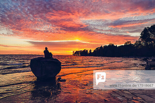 Woman sitting on large rock near the water watching the sunset.