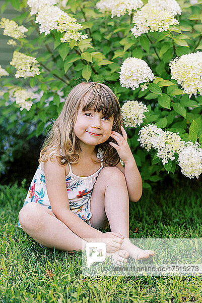 Little girl sitting on lawn in front of hydrangea bush
