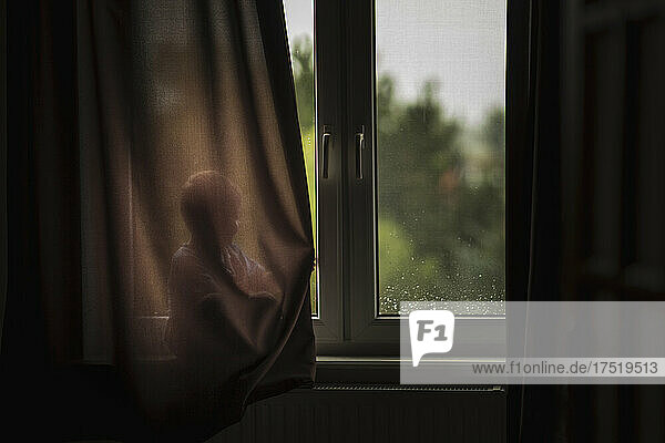 Child sitting on windowsill and hiding behind brown curtain