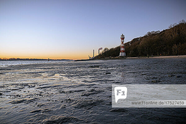 Eisiger Winternachmittag am Strand von Wittenbergen in Hamburg-Rissen. Die untergehende Sonne beleuchtet den Leuchtturm. Im Hintergrund die Schornsteine des Kohlekraftwerkes Wedel
