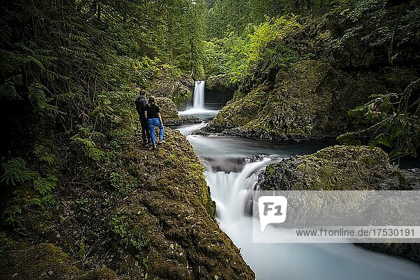 Paar steht am Fluss  Spirit Falls  Wasserfall fließt über Felsvorsprung  Basaltfelsen  Langzeitaufnahme  herbstlicher dichter Wald  Washington  USA  Nordamerika