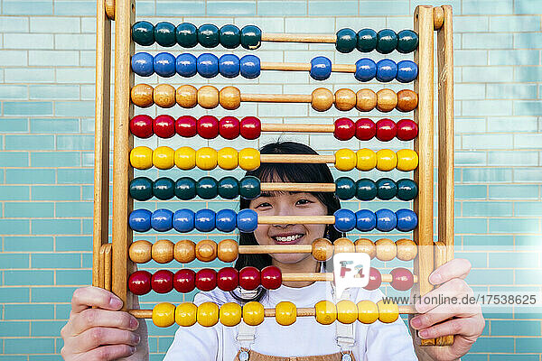 Smiling young woman holding abacus in front of brick wall