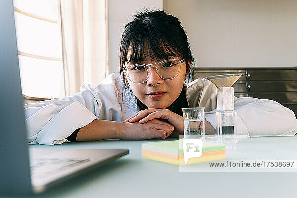 Young chemist wearing eyeglasses leaning on table in laboratory
