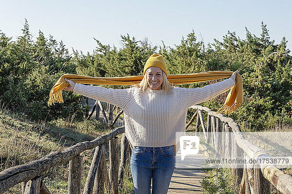 Smiling woman with arms outstretched holding scarf on wooden bridge