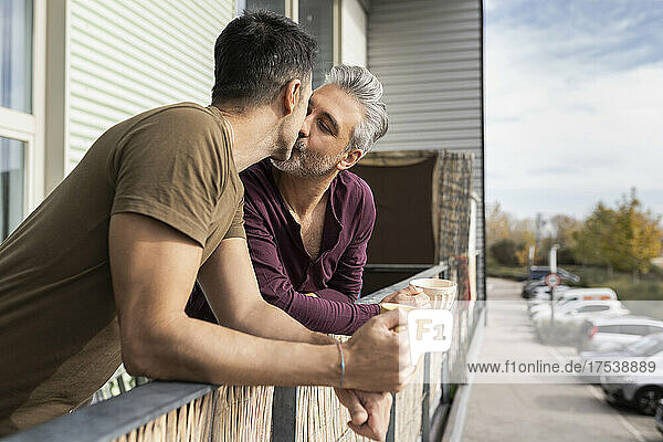 Romantic gay couple holding coffee mugs kissing in balcony