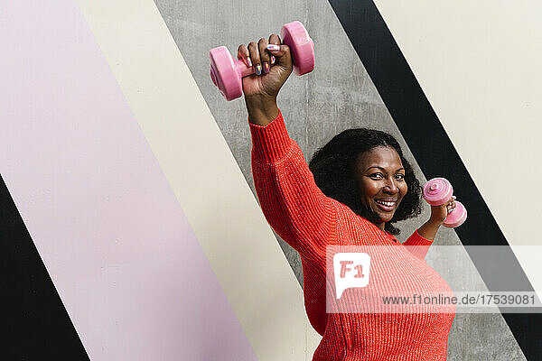 Happy plus size woman exercising with dumbbells near patterned wall