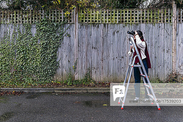 Young woman looking through binoculars standing by ladder on footpath