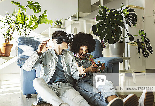 Young man with VR glasses gesturing by cheerful girlfriend playing video game at home