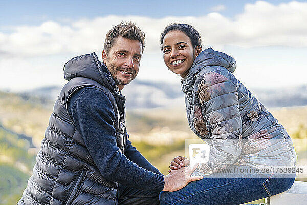 Smiling couple wearing padded jackets sitting at park