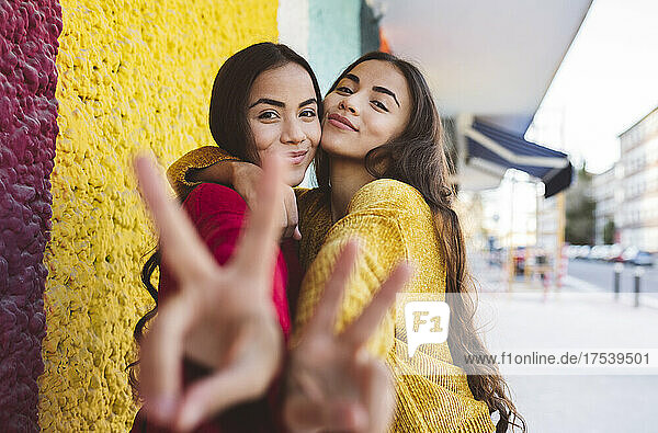 Young sisters embracing and gesturing with peace sign by colorful wall