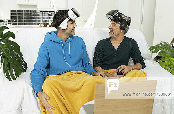 Smiling gay couple wearing VR glasses looking at each other sitting on sofa