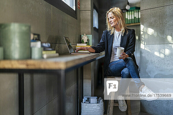 Businesswoman with coffee mug using laptop sitting on stool at office