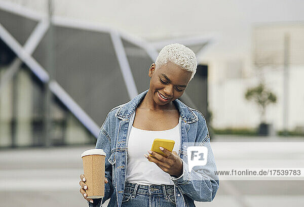 Smiling woman using smart phone holding disposable cup