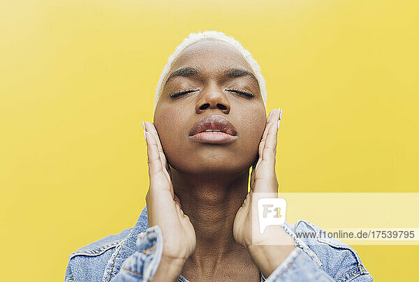 Young woman with eyes closed touching cheeks against yellow background