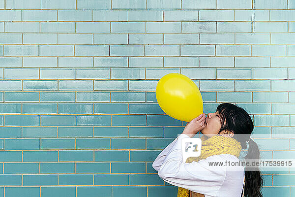 Woman wearing knitted scarf blowing yellow balloon by turquoise brick wall