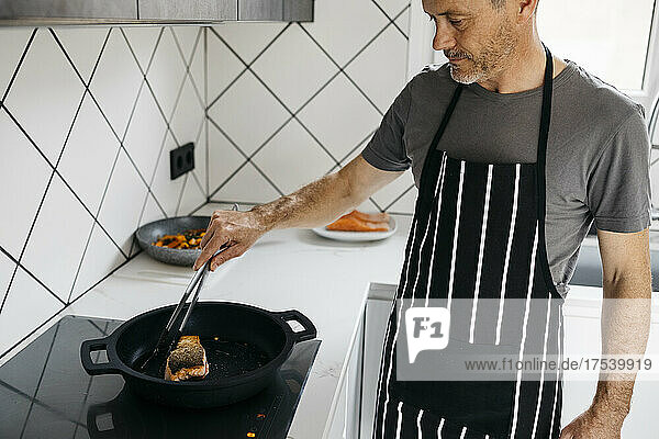 Man holding tongs checking cooked fish in pan at home