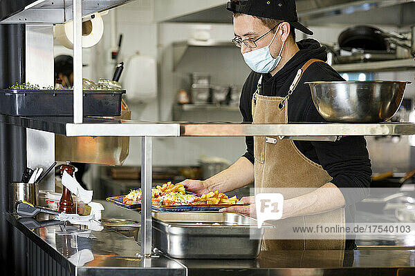 Chef with protective face mask holding food plates at restaurant kitchen