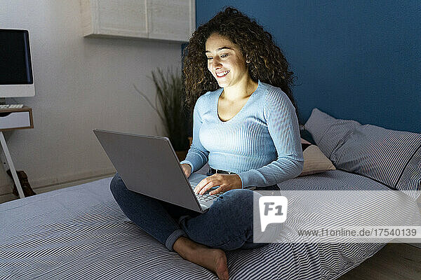 Junge Frau sitzt mit Laptop zu Hause im Bett
