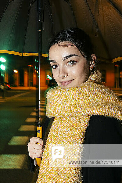 Smiling teenage girl with scarf and umbrella on street at night