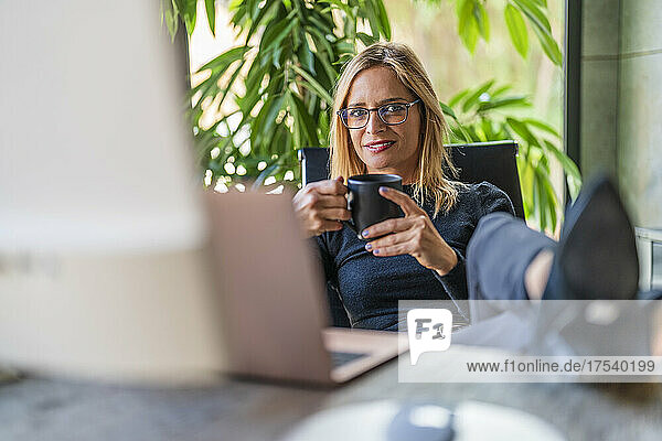 Geschäftsfrau hält Kaffeetasse in der Hand und sitzt mit erhobenen Füßen am Schreibtisch