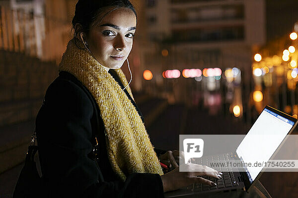 Teenage girl with laptop listening music on in-ear headphones at night