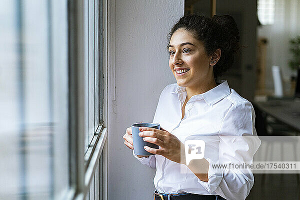 Smiling woman with mug looking out of window at home